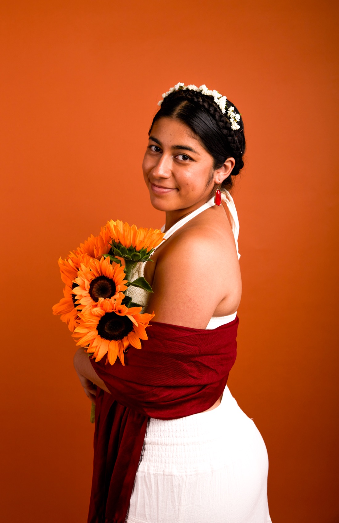 Metztli holding a bouquet of sunflowers, in a flower crown and red rebozo against a warm terracotta backdrop